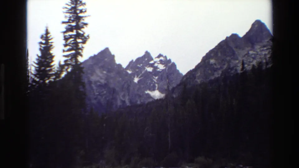 1980 Grand Teton home-movie frame with snow-marked peaks, dark forest, tall evergreens, and faded blue-purple color.