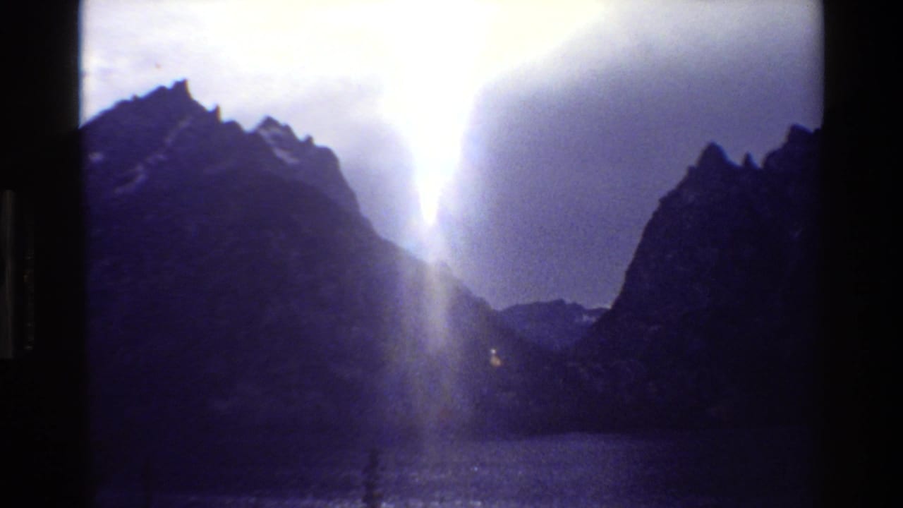 1980 8mm Grand Teton view of dark peaks around a lake as a bright shaft of sunlight breaks through heavy cloud.