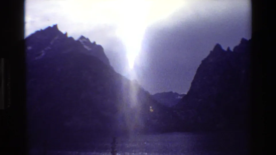 1980 8mm Grand Teton view of dark peaks around a lake as a bright shaft of sunlight breaks through heavy cloud.