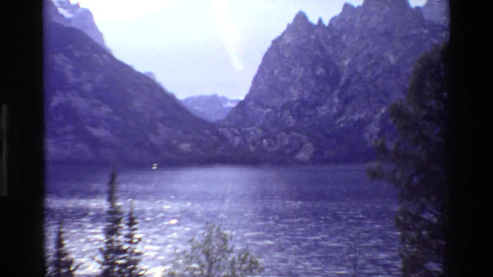 1980 Grand Teton home-movie frame with evergreens in the foreground, a bright lake, and mountain walls beyond.