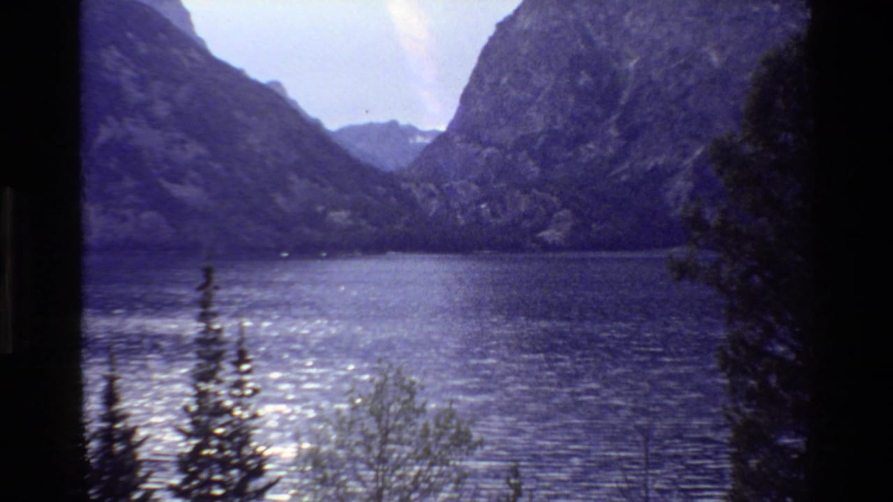 1980 Grand Teton home-movie frame showing a calm mountain lake below rugged slopes with pine trees in the foreground.