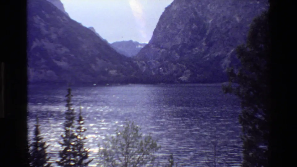1980 Grand Teton home-movie frame showing a calm mountain lake below rugged slopes with pine trees in the foreground.