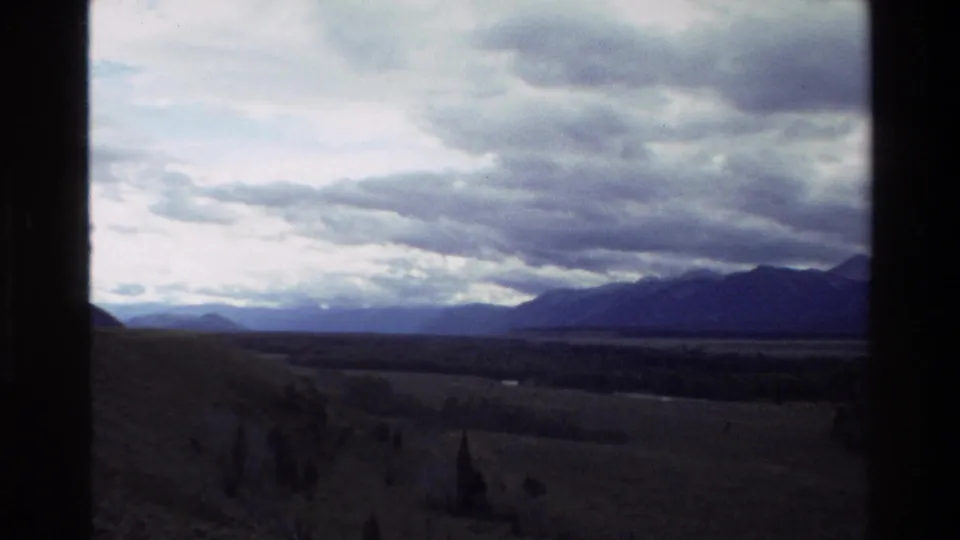 Grand Teton area valley view from 1980 8mm footage, with distant mountains under dramatic cloud cover.