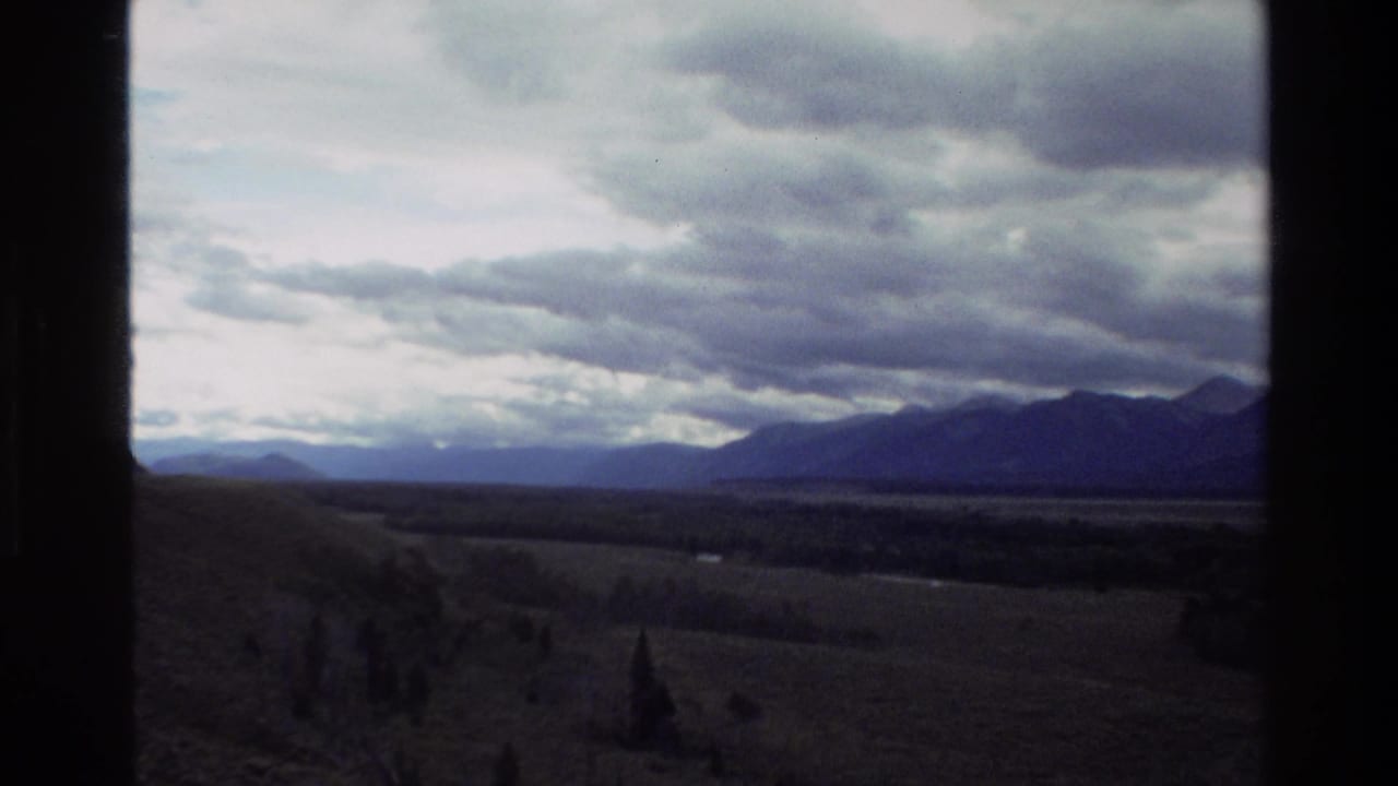 1980 8mm Grand Teton travel view showing cloudy mountains, open pasture, and a distant valley through dark window framing.