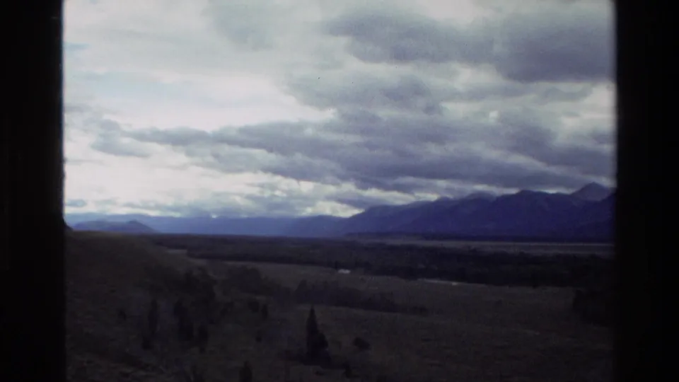 1980 8mm Grand Teton travel view showing cloudy mountains, open pasture, and a distant valley through dark window framing.