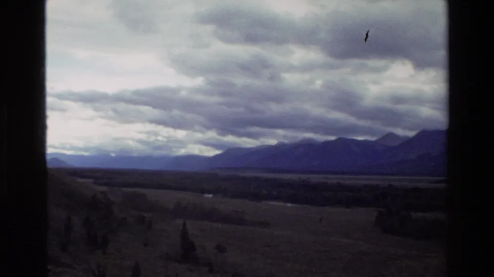 Grand Teton, Wyoming 8mm frame with layered mountains, a dim valley, pastureland, and heavy gray clouds seen from a moving vehicle.