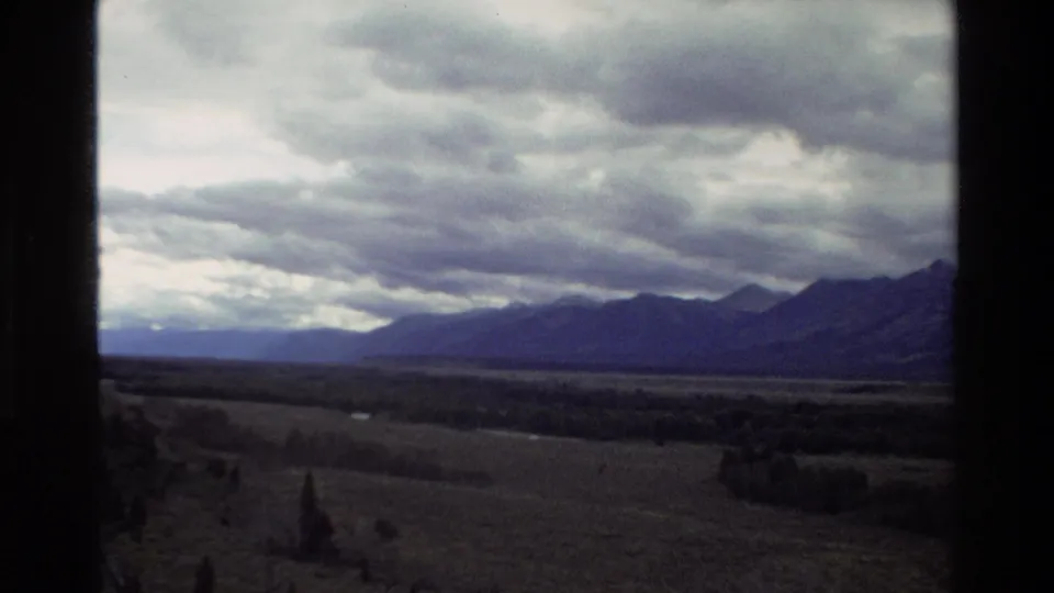 1980 home-movie frame of Grand Teton pastureland, distant mountain silhouettes, and layered clouds with soft 8mm color and window edges.