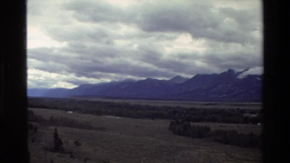Grand Teton 8mm travel frame showing pasture, a distant treeline, mountain ridges, and dramatic cloud cover through vehicle-window framing.
