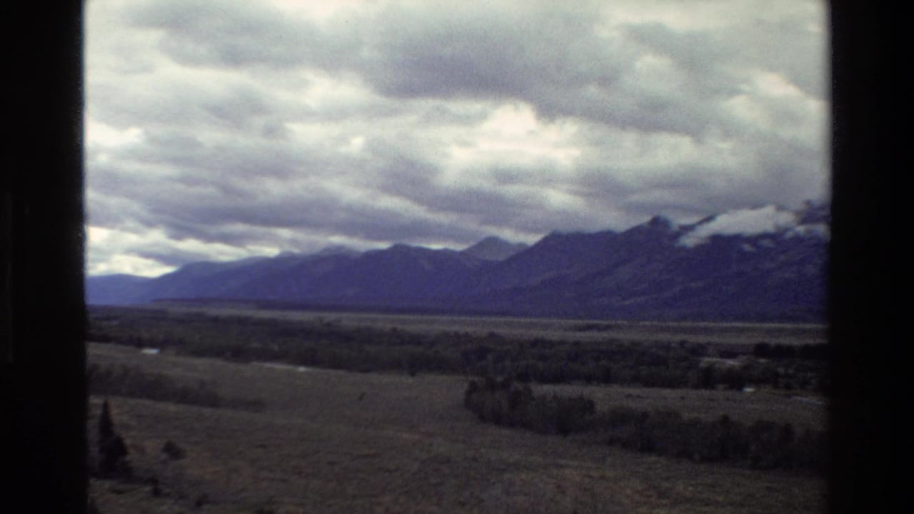 1980 8mm view of the Grand Teton range in Wyoming across a broad valley under heavy clouds, with dark vertical frame edges.