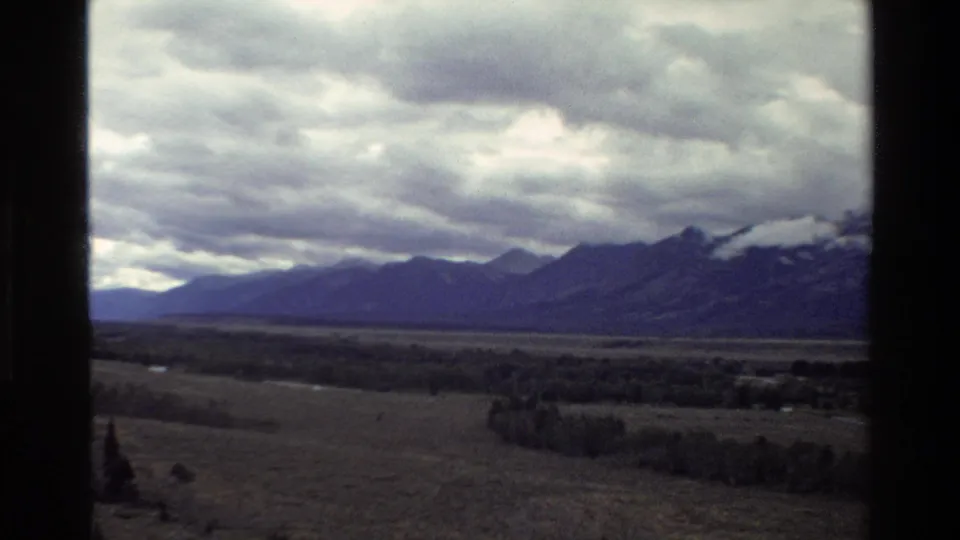 1980 8mm view of the Grand Teton range in Wyoming across a broad valley under heavy clouds, with dark vertical frame edges.