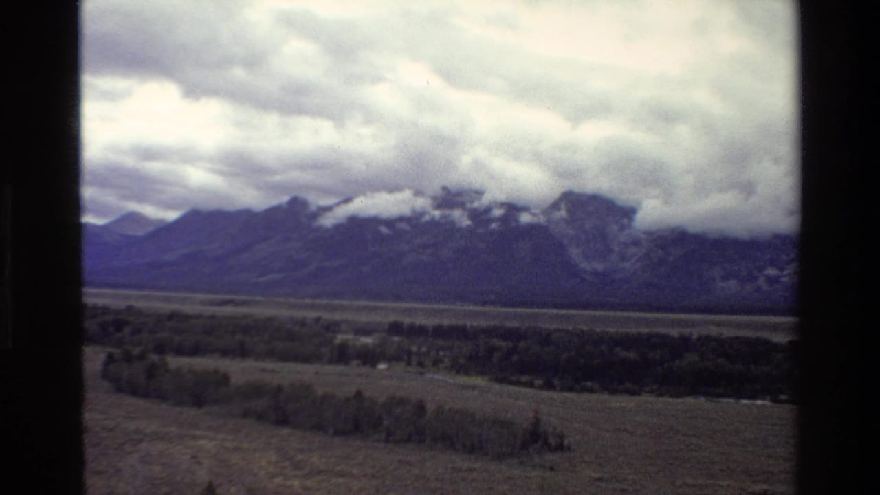 1980 8mm Grand Teton view showing the Teton Range beneath low clouds, with open valley and scattered trees in the foreground.