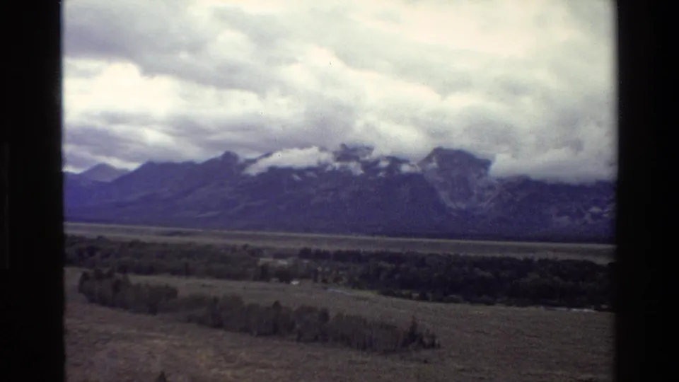 1980 8mm Grand Teton view showing the Teton Range beneath low clouds, with open valley and scattered trees in the foreground.