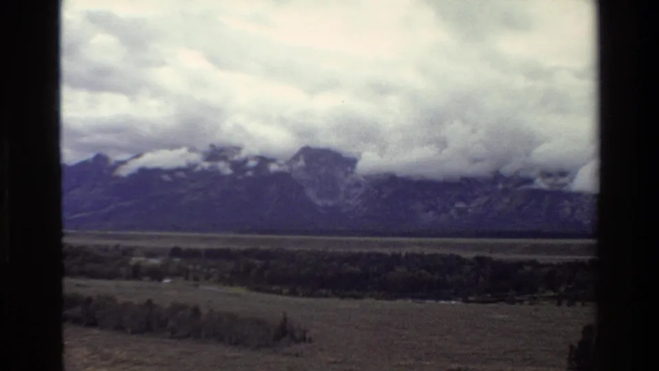 Soft 1980 home-movie frame of the Teton Range under dense clouds, looking across trees and open valley land.
