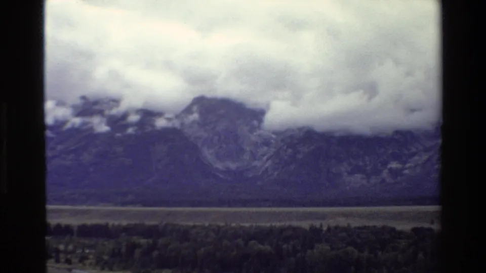 Vintage 8mm Grand Teton view with purple-blue color shift, storm clouds over mountains, and a dark tree line below.