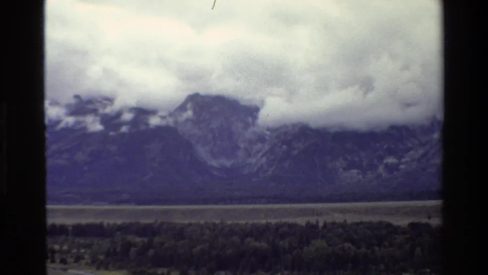 1980 Grand Teton home-movie frame showing rugged mountains partly covered by low cloud above a dark forest and valley floor.