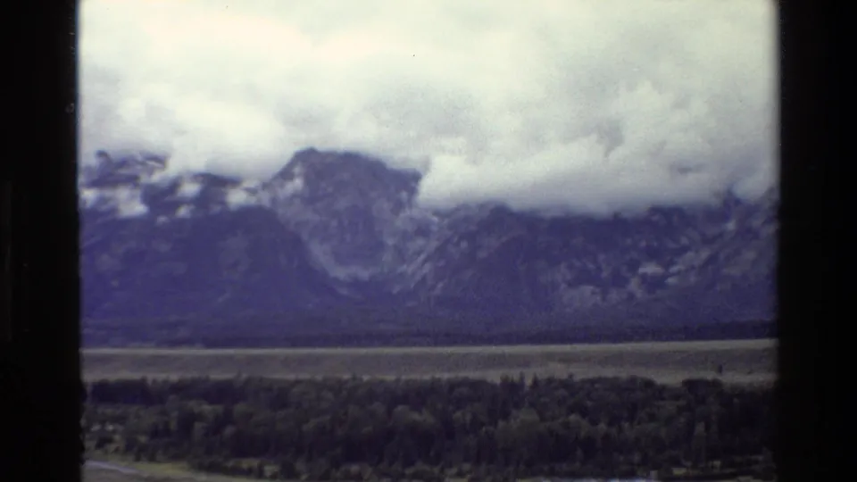 Vintage 8mm view of the Grand Teton range with blue-purple color shift, low clouds, forest, and a pale strip of valley below.
