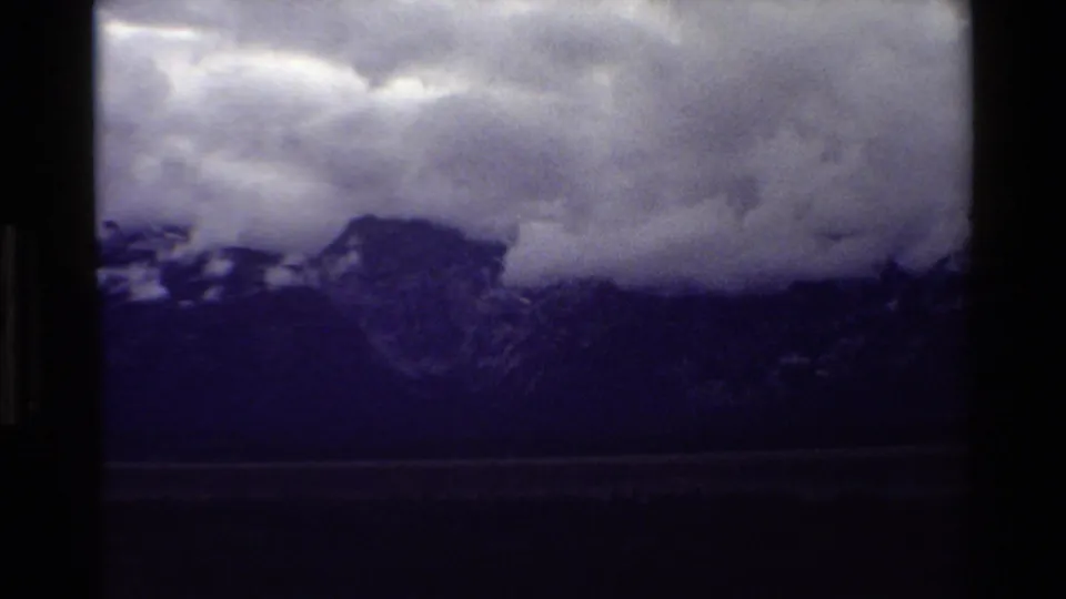 Darker 8mm frame from Grand Teton showing the mountain silhouette under dense clouds with a dim valley and forested foreground.