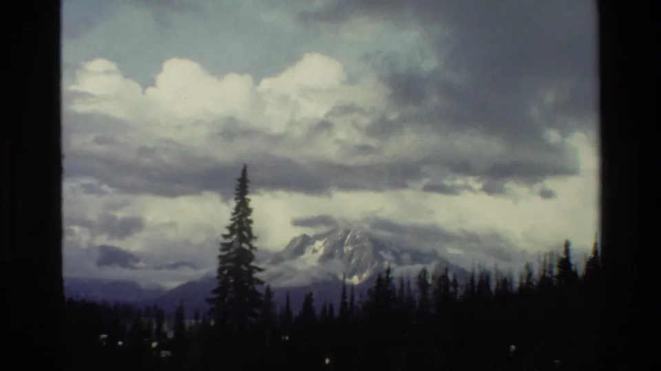 1980 Grand Teton, Wyoming frame with a snowy mountain slope, dark forest silhouettes, and thick gray clouds across the sky.
