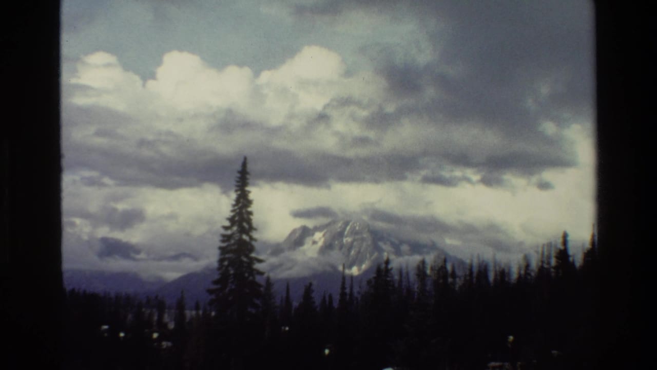Grand Teton mountain view in Wyoming, 1980, with snow-covered peaks partly obscured by clouds and a dark evergreen treeline in the foreground.