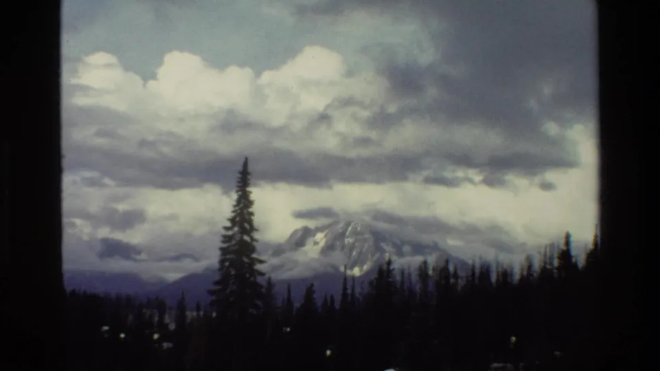 Grand Teton mountain view in Wyoming, 1980, with snow-covered peaks partly obscured by clouds and a dark evergreen treeline in the foreground.