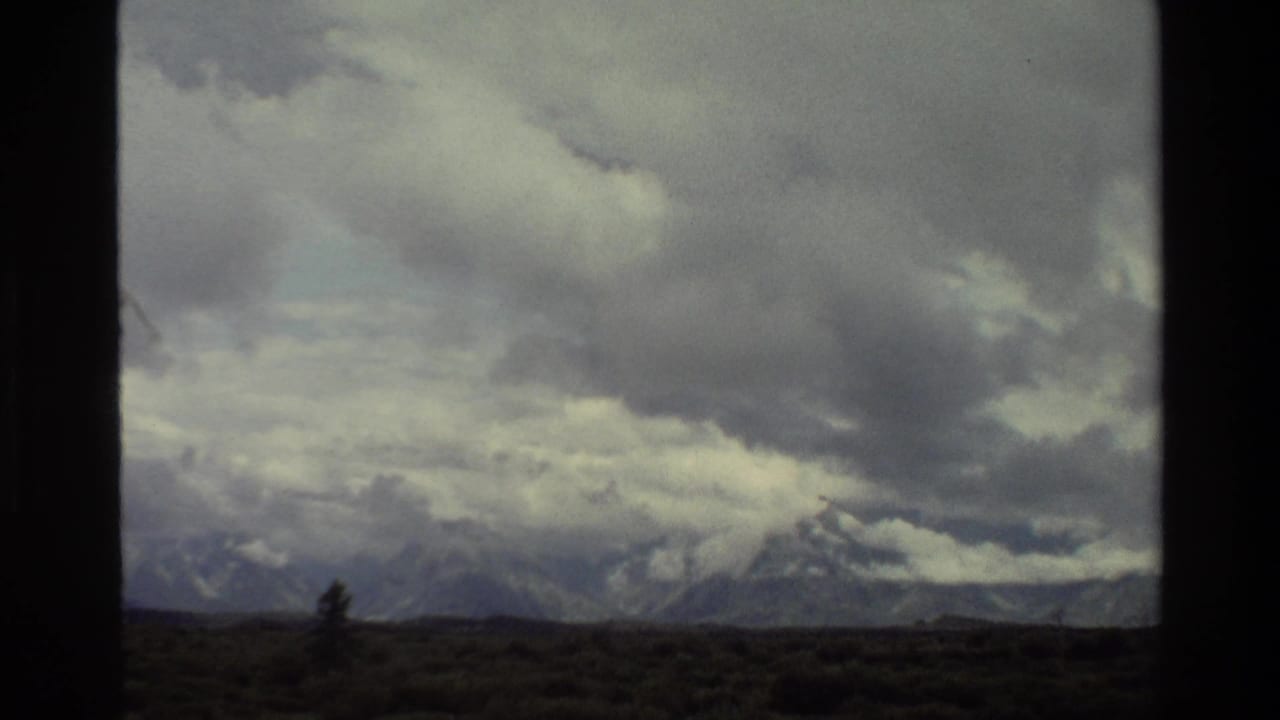1980 8mm view of the Grand Teton range under heavy clouds, seen across dark pasture with a window-like divider in the frame.