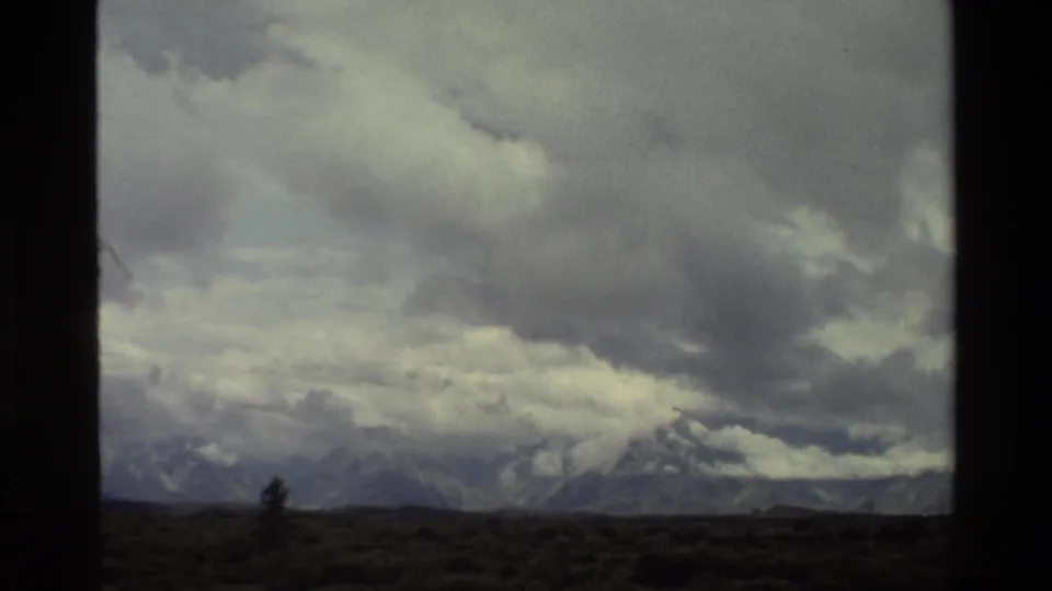 1980 8mm view of the Grand Teton range under heavy clouds, seen across dark pasture with a window-like divider in the frame.