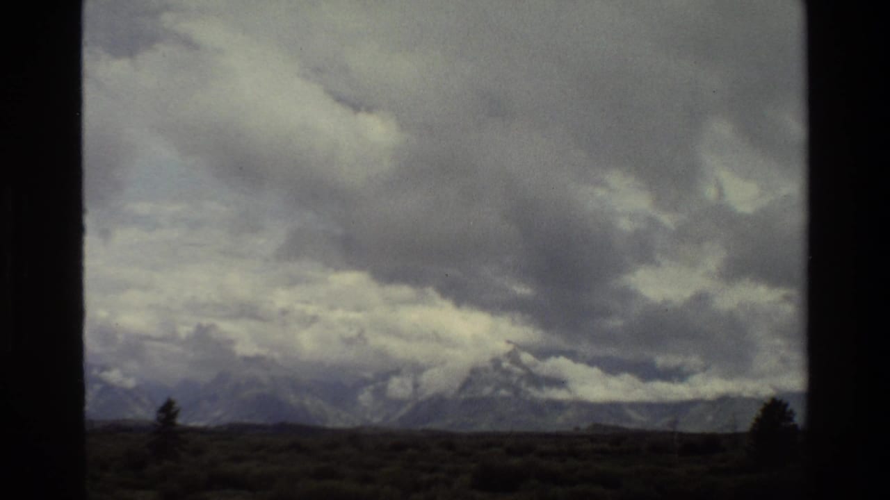 Grand Teton landscape in 1980 home-movie footage, with snow-streaked peaks, low clouds, and a dark foreground plain.