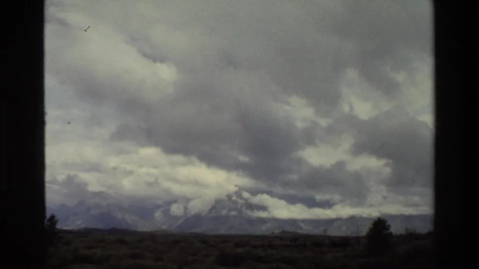 1980 8mm frame of the Grand Teton mountains partly obscured by clouds, with scrubby foreground and a lone dark conifer.