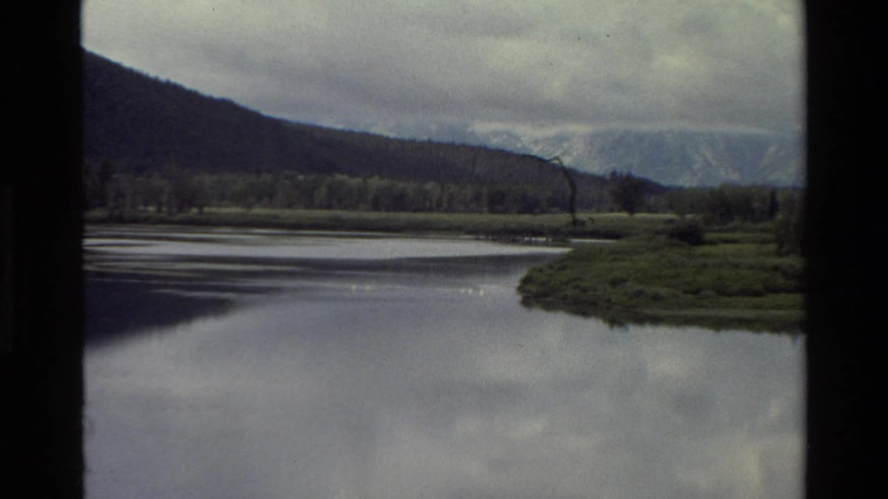 1980 Grand Teton home-movie frame showing a calm river bend, green valley edges, forested slopes, clouds, and distant mountains.