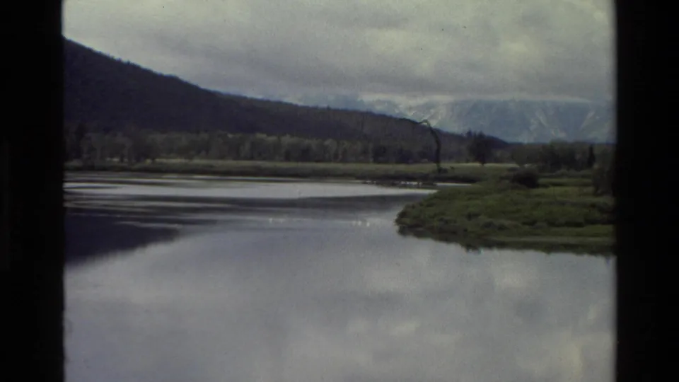 1980 Grand Teton home-movie frame showing a calm river bend, green valley edges, forested slopes, clouds, and distant mountains.