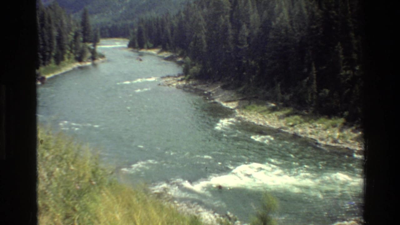 Grand Teton river scene showing evergreen banks, rocky shoreline, and white-water forming in the lower frame.
