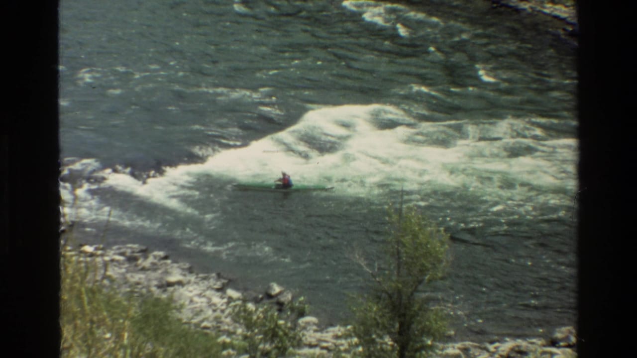 1980 Grand Teton home-movie frame showing a distant kayaker in a green kayak moving toward foaming rapids on a mountain river.