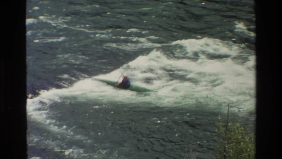Closer 1980 Grand Teton frame showing a helmeted kayaker in a green kayak riding through whitewater rapids.