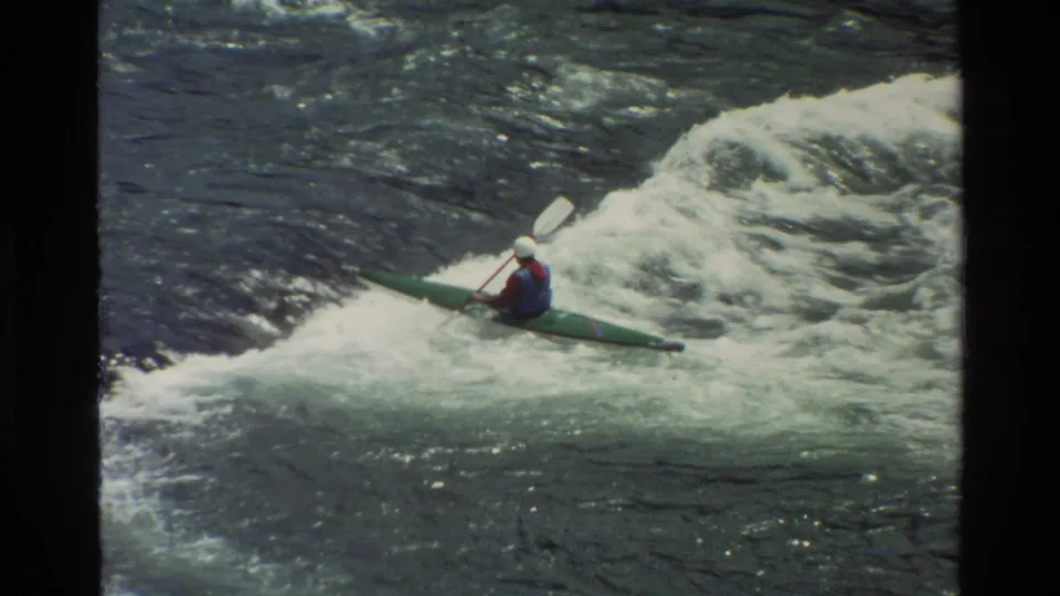 1980 Grand Teton home-movie frame of a kayaker in a green boat using a white paddle beside churning whitewater.