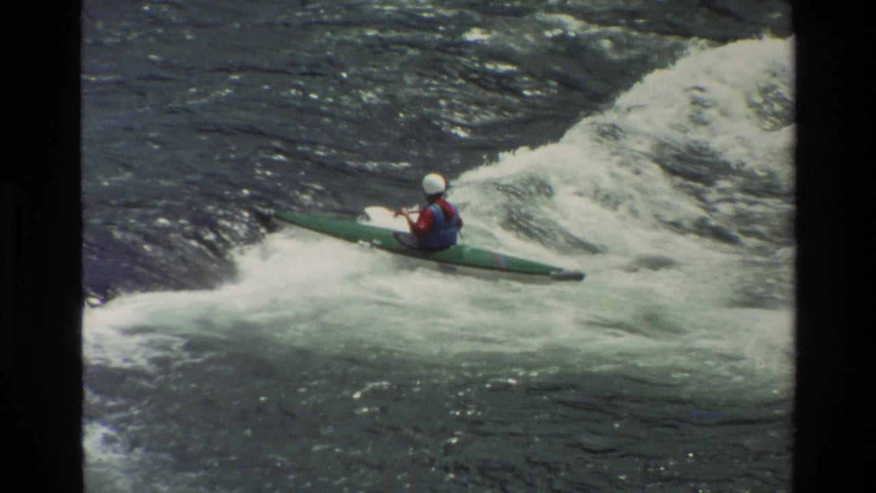 1980 Grand Teton, Wyoming home-movie frame showing a paddler in a green boat wearing a white helmet and red vest in rapids.