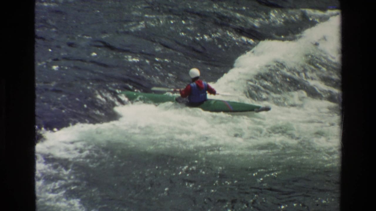 Paddler in a green boat moving across foaming river rapids.