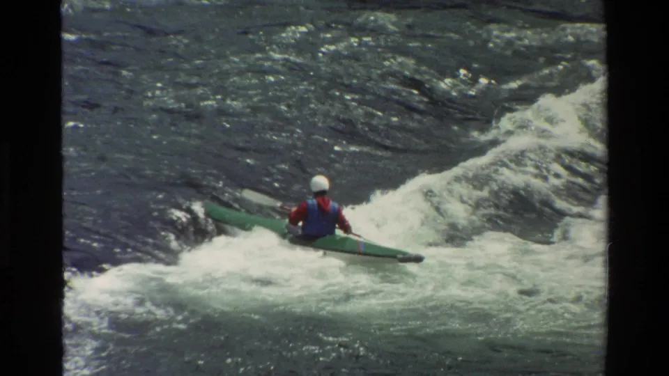 1980 Grand Teton, Wyoming frame showing a distant helmeted paddler in a green boat among churning rapids.