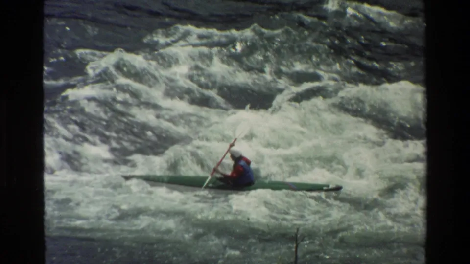 Side view of a helmeted kayaker paddling a green kayak through churning whitewater in the 1980 Wyoming clip.