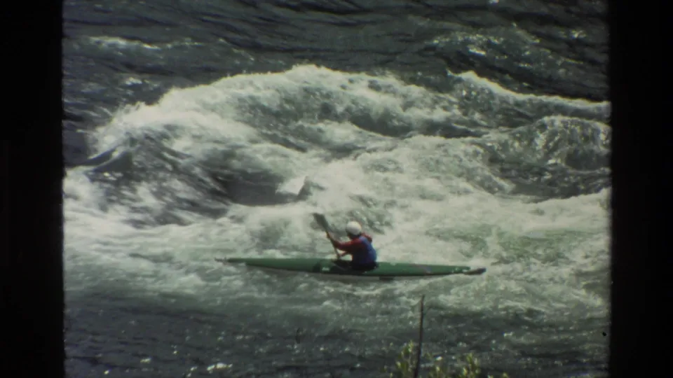 A helmeted kayaker in a green kayak continues through foaming rapids in archival Grand Teton, Wyoming footage.