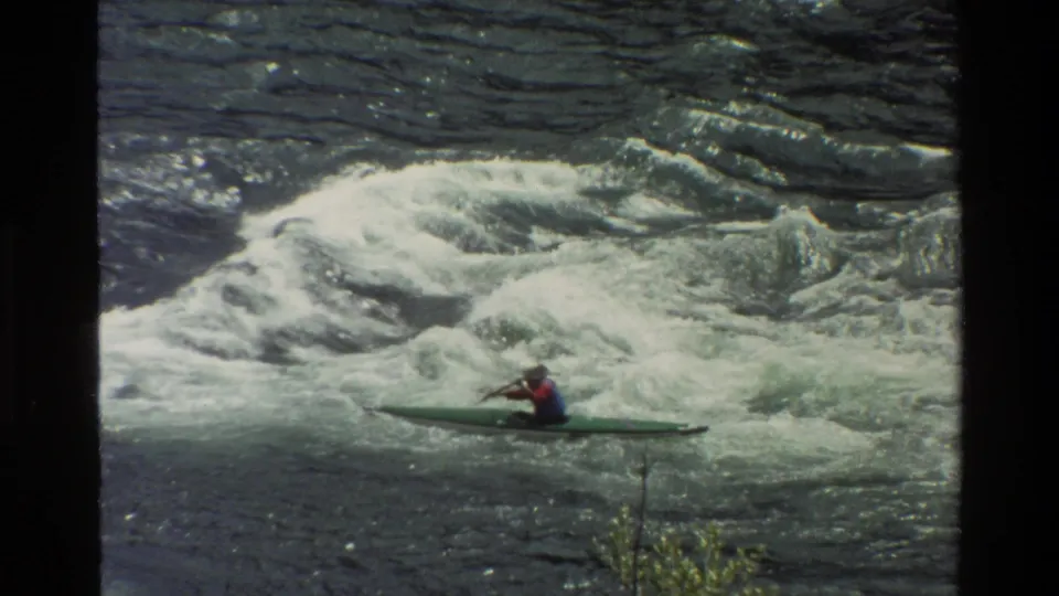 A helmeted kayaker in a green kayak moves through churning rapids in Grand Teton, Wyoming, in 1980 8mm footage.