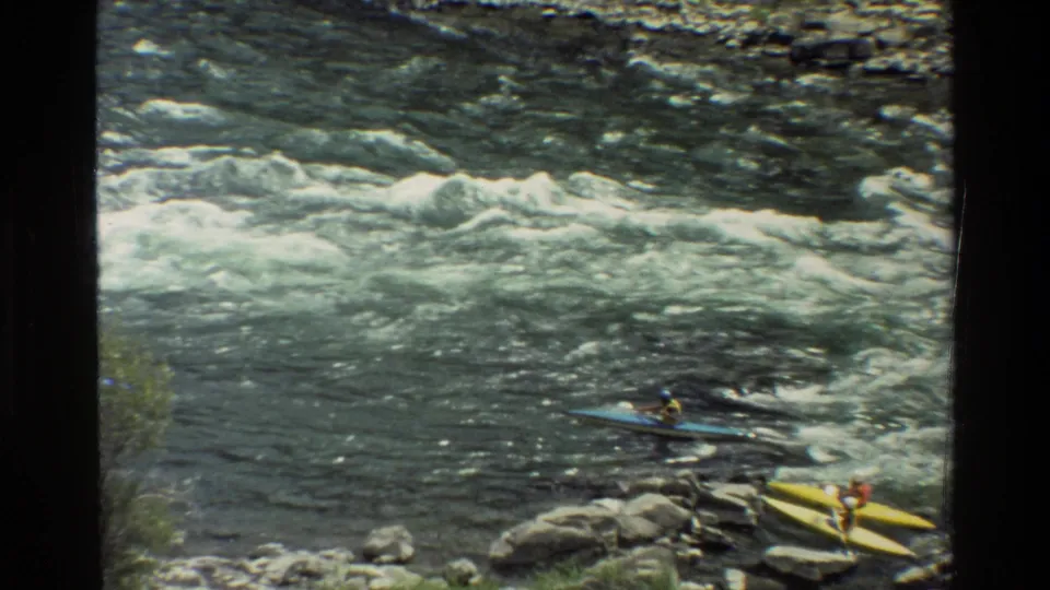 From a high angle, blue and yellow kayaks sit near rocks while rapids run across the upper part of the frame.