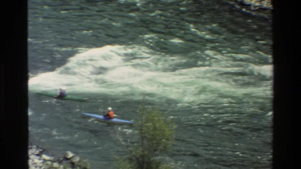 Two kayakers, including one in a blue boat and one in a green boat, paddle below a foaming rapid in vintage 8mm footage.
