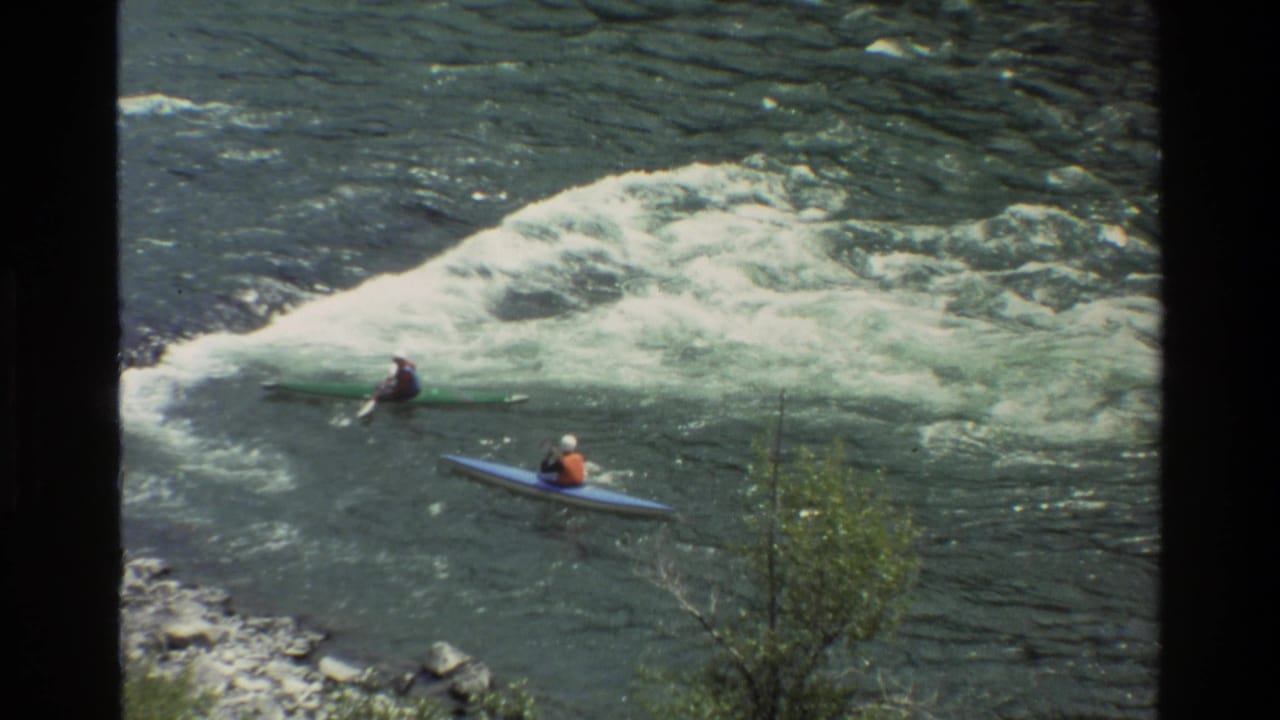1980 home-movie frame from Grand Teton, Wyoming showing two kayakers near churning river rapids, filmed from above the bank.