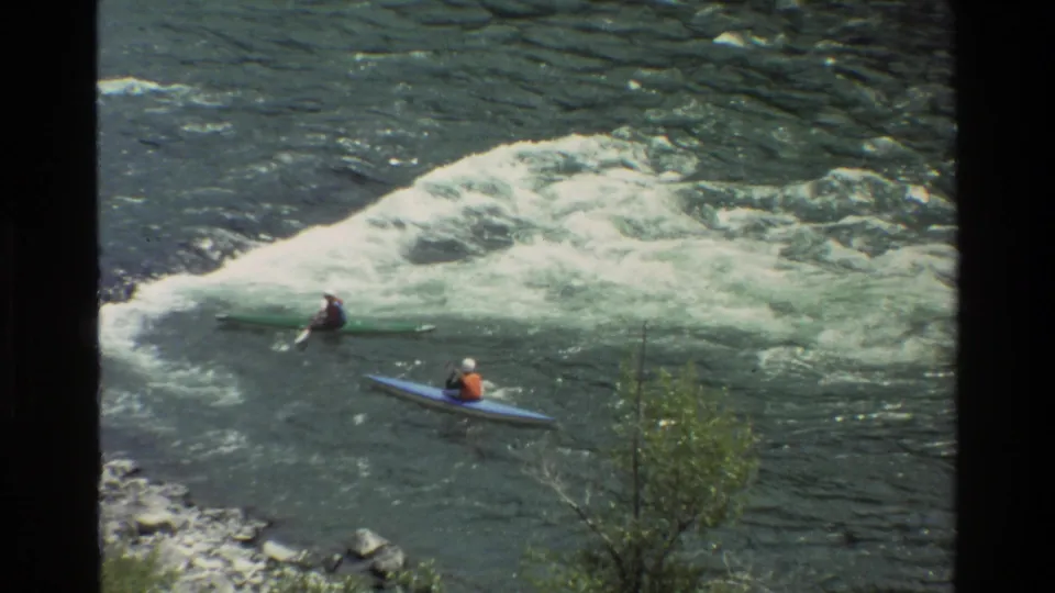 1980 home-movie frame from Grand Teton, Wyoming showing two kayakers near churning river rapids, filmed from above the bank.