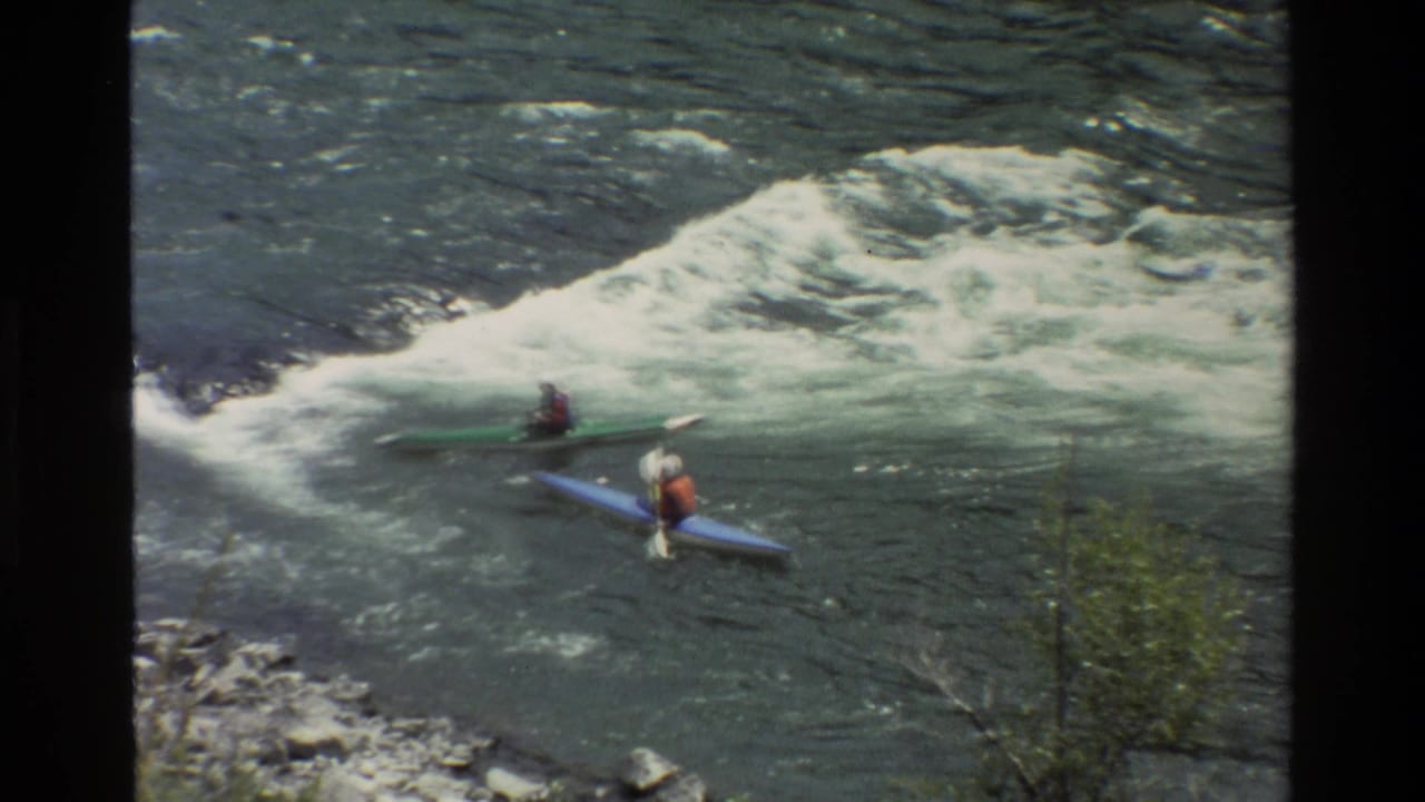 Two kayakers paddle near a foaming rapid while one boat turns across the current.