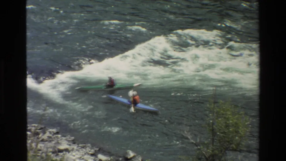 Grand Teton river footage shows kayakers in blue and green boats maneuvering beside whitewater, with brush and rocks at the lower edge.