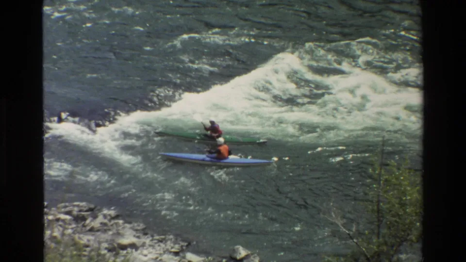 A handheld 8mm frame shows two river paddlers below foaming rapids, one in a blue kayak and one in a green kayak.