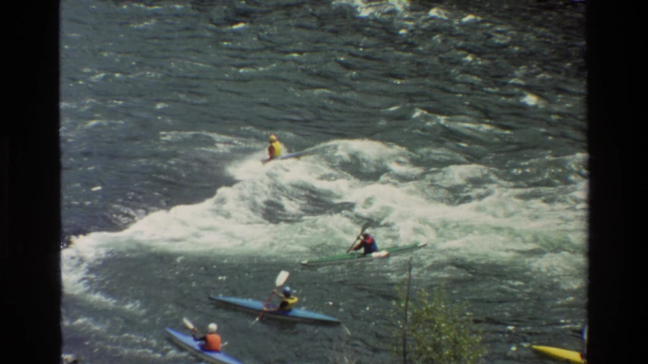 Kayakers spread across a foamy rapid with one paddler near the wave crest.