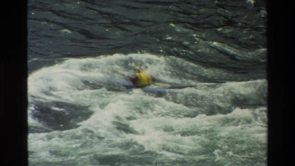 Close 1980 home-movie frame from Grand Teton showing a kayaker in a yellow vest cutting through rough whitewater.