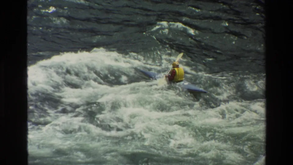 1980 Grand Teton home-movie frame showing a kayaker in a yellow life jacket navigating foamy whitewater.
